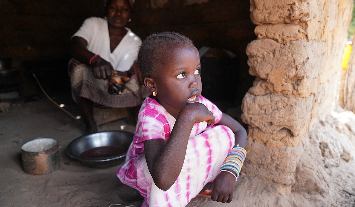 A young girl sits on the floor of a mud hut.