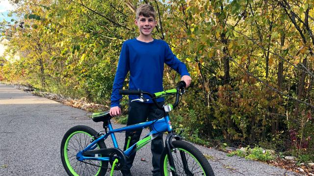 A boy with a blue and green bike standing next to a tree.