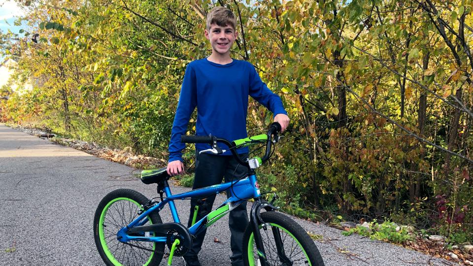 A boy with a blue bike standing next to a tree.