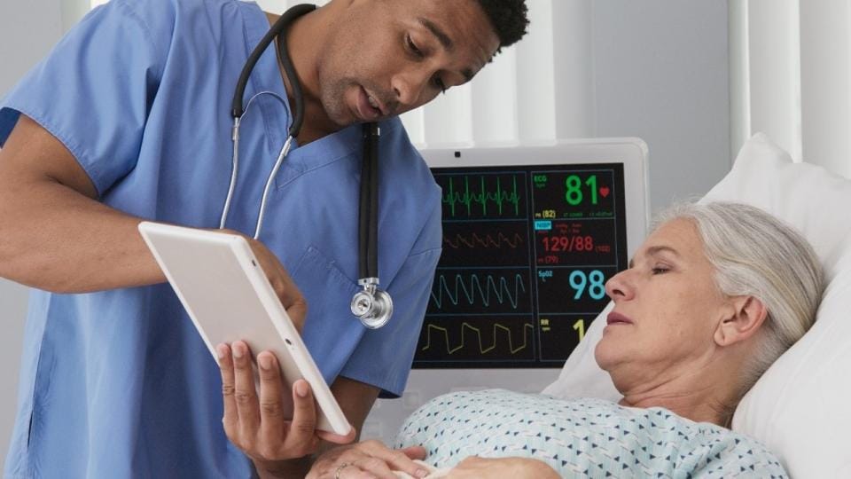 A nurse is looking at a tablet while an older woman is laying on a bed.