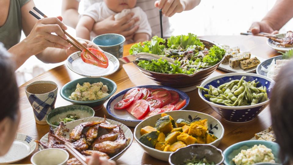 A table filled with various dishes, including salad, edamame, sliced tomatoes, grilled fish, and squash, with people using chopsticks and a baby held in the background.