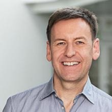 A man in a gray shirt smiles in front of a building.