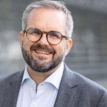 A man with glasses and a beard smiles in front of a building.