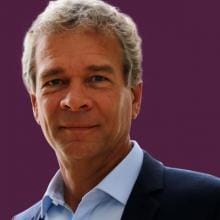 A man in a suit stands in front of a purple background.