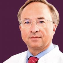 A man in a lab coat and tie poses against a purple background.