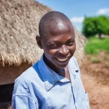 A boy smiles in front of a thatched hut.