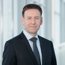 A man in a business suit stands in front of a glass wall.