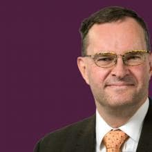 A man in a suit and tie stands in front of a purple background.