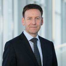 A man in a business suit stands in front of a glass wall.