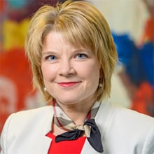 A woman in a white jacket stands in front of a painting.