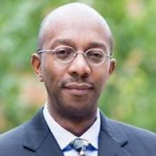 A black man in a suit and tie stands in front of a tree.