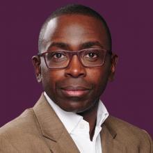 A man in glasses is standing in front of a purple background.