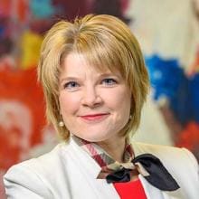 A woman in a white blazer and red shirt standing in front of a painting.