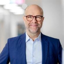 A business man, with glasses and a blue suit, smiles into the camera.